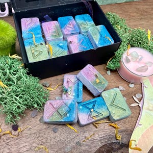 May include: A collection of rectangular resin tiles featuring gradient blue, pink, and green colors with engraved runic symbols. Some tiles are housed in a black box, while others are scattered alongside a small pink candle and a moth cutout.