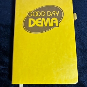 Peut inclure: Un cahier jaune avec le texte "GOOD DAY DEMA" dans un ovale marron. Le cahier a un signet en ruban et une couverture texturée. Le fond est un tissu bleu foncé.