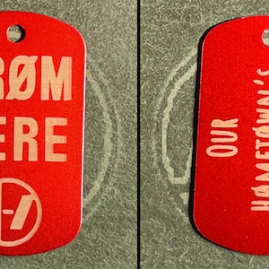 May include: A red rectangular metal dog tag with rounded corners and a hole at the top. The front features the text FRØM HERE and a circular logo with a slash. The back features the text OUR HØMETØWN'S IN THE DARK in a sans-serif font.