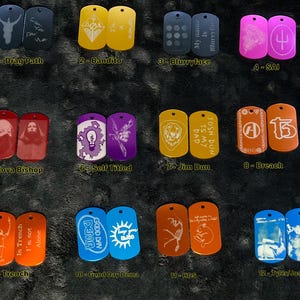 Puede incluir: Una colección de doce pares de chapas grabadas en varios colores. Cada chapa presenta diseños y textos únicos, incluyendo ilustraciones, símbolos y frases como "Drag Path" y "Blurryface". Las chapas están dispuestas sobre una superficie oscura y texturizada.