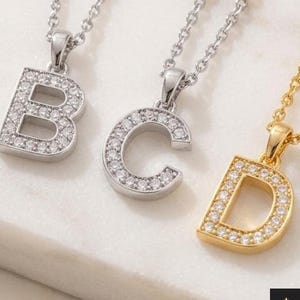Puede incluir: Cuatro collares con colgantes de letras A, B, C y D. Los colgantes A y D son de tono dorado con piedras claras, mientras que los colgantes B y C son de tono plateado con piedras claras. Cada colgante cuelga de una fina cadena de metal.