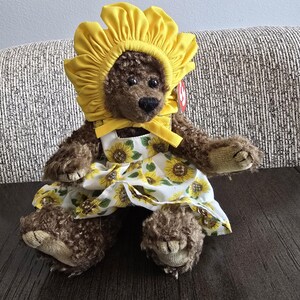 Könnte beinhalten: Ein brauner Teddybär mit einer gelben Sonnenblumenhaube und einem weißen Kleid mit Sonnenblumenmuster. Der Bär sitzt auf einer dunkelbraunen Oberfläche. Das Kleid hat gelbe Träger und eine gelbe Schleife.