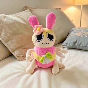 Puede incluir: Un peluche de conejito con orejas rosas, cuerpo amarillo y bufanda rosa. El conejito tiene una expresión triste con ojos negros y una pequeña flor en la cabeza. Sostiene un pequeño objeto verde y blanco.