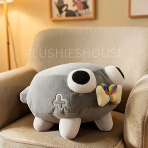 Puede incluir: Un peluche gris con ojos grandes blancos y negros, un peque&ntilde;o lazo y un dise&ntilde;o de tr&eacute;bol de cuatro hojas. El juguete est&aacute; sobre un sill&oacute;n beige. El texto "PLUSHIESHOUSE" es visible.