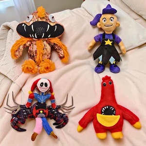 May include: Four handmade plush toys in a variety of monster and character designs. The toys include an orange monster with a toothy grin, a witch doll, a clown-like figure with crab-like arms, and a red creature with a yellow pouch.