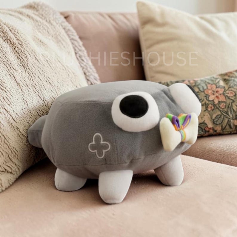 Puede incluir: Un peluche gris con forma de criatura con grandes ojos blancos y negros, pies blancos y un peque&ntilde;o lazo blanco con detalles de arco&iacute;ris. Un dise&ntilde;o en forma de cruz est&aacute; en el lateral. La palabra "SHIESHOUSE" es visible en el fondo.