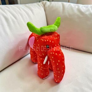 Puede incluir: Un elefante de peluche con forma de fresa. El cuerpo rojo está salpicado de semillas blancas, con orejas rosas y colmillos blancos. La parte superior presenta una hoja verde. El texto "PLUSHIESHOUSE" es visible en el lateral.