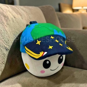 Puede incluir: Un peluche con forma de luna sonriente que lleva una gorra de béisbol. La gorra presenta un diseño de la Tierra verde y azul en la parte superior y un cielo estrellado en la visera. El juguete tiene un cuerpo blanco, mejillas rosas y el texto "PLUSHIESHOUSE".