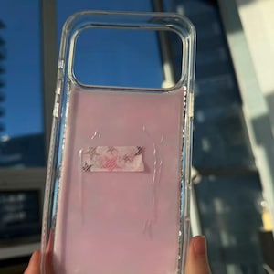 May include: Clear phone case with a pink interior and a decorative tape strip. The case is held up against a window with a blue sky visible in the background.