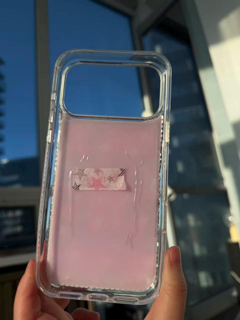 May include: Clear phone case with a pink interior and a decorative tape strip. The case is held up against a window with a blue sky visible in the background.