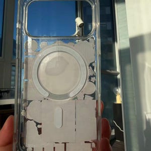 Pu&ograve; includere: Custodia protettiva trasparente per telefono con anello MagSafe bianco e residui adesivi bianchi. La custodia ha una forma rettangolare con angoli arrotondati e ritagli per la fotocamera e i pulsanti. La custodia &egrave; tenuta in mano.