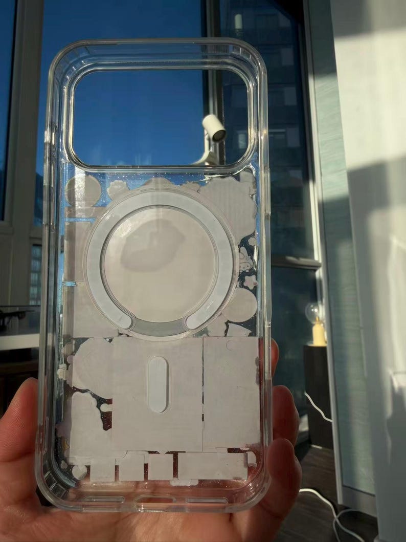 Pu&ograve; includere: Custodia protettiva trasparente per telefono con anello MagSafe bianco e residui adesivi bianchi. La custodia ha una forma rettangolare con angoli arrotondati e ritagli per la fotocamera e i pulsanti. La custodia &egrave; tenuta in mano.