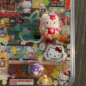 Op de afbeelding: Een doorzichtig telefoonhoesje versierd met een collage van Sanrio-karakterstickers, waaronder Hello Kitty, en kleine harsbedels met een 3D Hello Kitty-figuurtje, een wit Hello Kitty-gezicht en een paarse paddenstoel met witte stippen.
