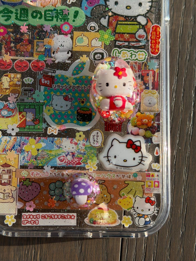 Op de afbeelding: Een doorzichtig telefoonhoesje versierd met een collage van Sanrio-karakterstickers, waaronder Hello Kitty, en kleine harsbedels met een 3D Hello Kitty-figuurtje, een wit Hello Kitty-gezicht en een paarse paddenstoel met witte stippen.