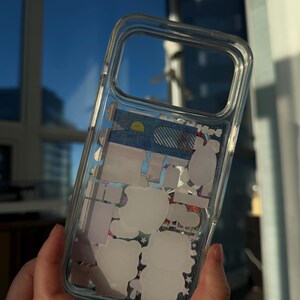 Puede incluir: Funda transparente para tel&eacute;fono con pegatinas decorativas en el interior. La funda presenta pegatinas blancas de personajes de dibujos animados y otras formas. La funda se sostiene frente a una ventana con un cielo azul de fondo.