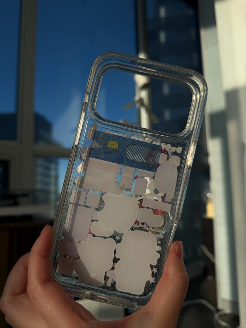 Puede incluir: Funda transparente para tel&eacute;fono con pegatinas decorativas en el interior. La funda presenta pegatinas blancas de personajes de dibujos animados y otras formas. La funda se sostiene frente a una ventana con un cielo azul de fondo.