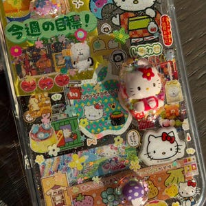 Op de afbeelding: Een doorzichtig telefoonhoesje versierd met een collage van Sanrio-karakterstickers, waaronder Hello Kitty en Pompompurin, samen met Japanse tekst, glitteraccenten en kleine 3D-harsbedels met een paddenstoel en een Hello Kitty-figuurtje.