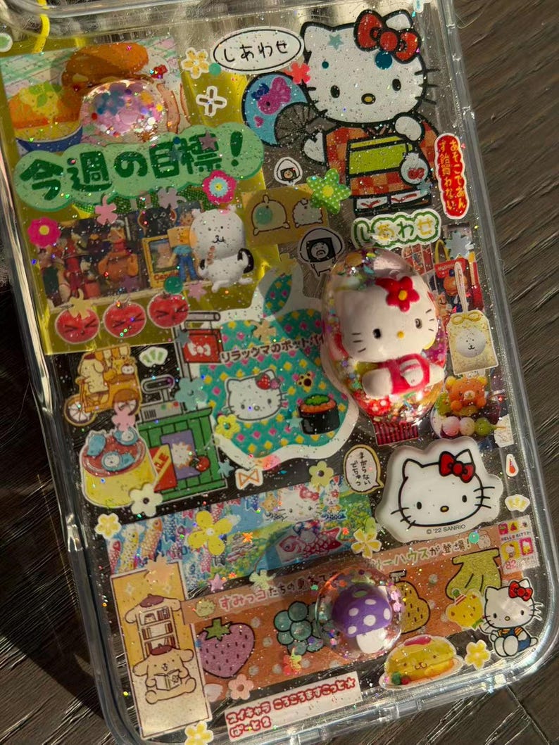 Op de afbeelding: Een doorzichtig telefoonhoesje versierd met een collage van Sanrio-karakterstickers, waaronder Hello Kitty en Pompompurin, samen met Japanse tekst, glitteraccenten en kleine 3D-harsbedels met een paddenstoel en een Hello Kitty-figuurtje.
