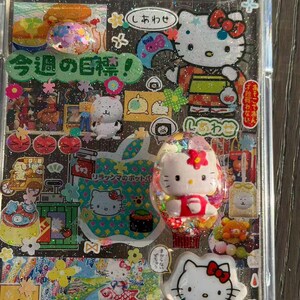 Op de afbeelding: Een doorzichtig telefoonhoesje versierd met een collage van Sanrio-karakterstickers, waaronder Hello Kitty, en diverse Japanse tekstelementen zoals '今週の目標'. Het ontwerp bevat glitteraccenten, kleurrijke bloemmotieven en kleine driedimensionale karakterbedels die op het oppervlak zijn bevestigd.