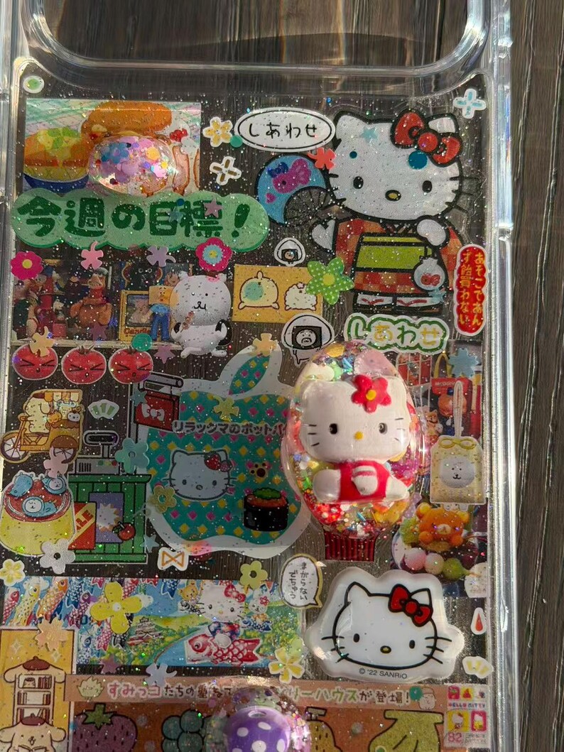Op de afbeelding: Een doorzichtig telefoonhoesje versierd met een collage van Sanrio-karakterstickers, waaronder Hello Kitty, en diverse Japanse tekstelementen zoals '今週の目標'. Het ontwerp bevat glitteraccenten, kleurrijke bloemmotieven en kleine driedimensionale karakterbedels die op het oppervlak zijn bevestigd.