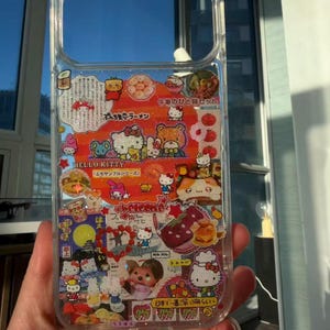 Pu&ograve; includere: Custodia trasparente per telefono con personaggi di Hello Kitty e testo giapponese. La custodia presenta immagini di Hello Kitty, cibo e altri elementi decorativi. La custodia &egrave; tenuta davanti a una finestra.