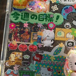 Op de afbeelding: Een doorzichtig telefoonhoesje versierd met diverse kleurrijke stickers, waaronder een groen lint met Japanse tekst, een witte beer, een Hello Kitty-afbeelding en kleine bloemvormige accenten, alles tegen een achtergrond met glitters.