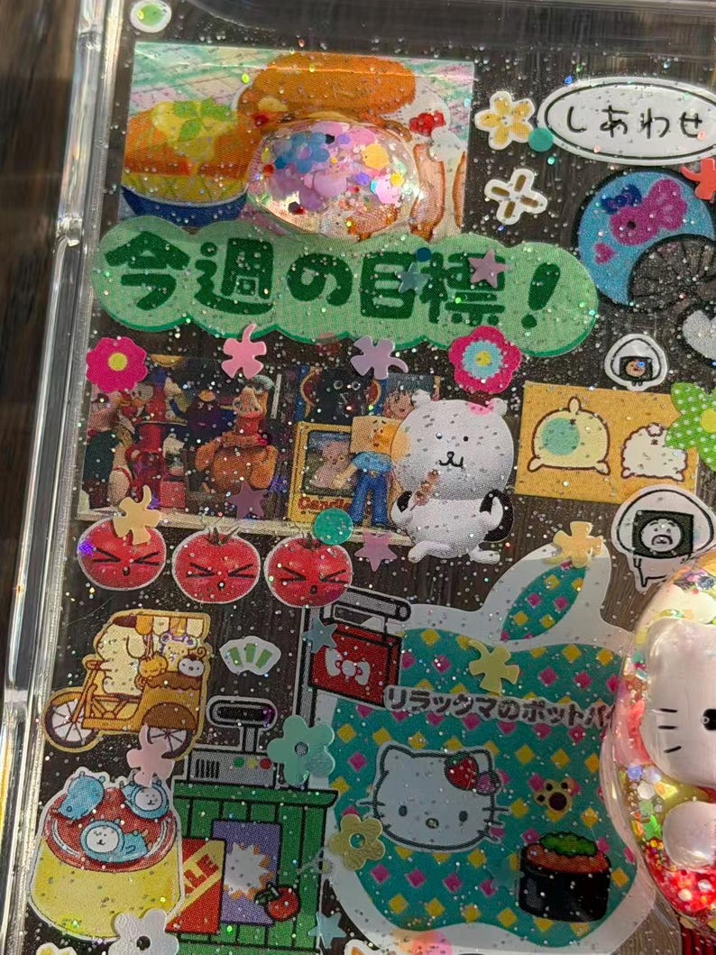 Op de afbeelding: Een doorzichtig telefoonhoesje versierd met diverse kleurrijke stickers, waaronder een groen lint met Japanse tekst, een witte beer, een Hello Kitty-afbeelding en kleine bloemvormige accenten, alles tegen een achtergrond met glitters.