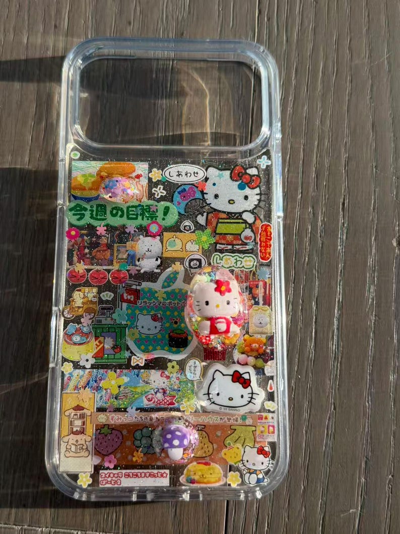 Op de afbeelding: Een doorzichtig telefoonhoesje versierd met een collage van kleurrijke stickers en driedimensionale bedels. Het ontwerp bevat meerdere Hello Kitty-illustraties, Japanse tekst waaronder "今週の目標!", diverse voedselpictogrammen en kleine glinsterende accenten die over het hele oppervlak zijn verwerkt.
