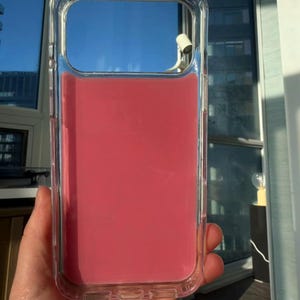 May include: Clear protective phone case with a vibrant pink interior. The case is held up against a bright blue sky, showcasing its transparent design and the color of the phone it protects. The case is rectangular with rounded corners.