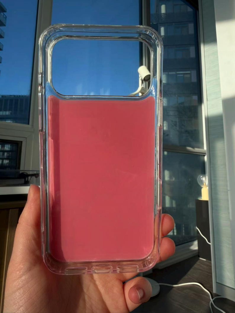 May include: Clear protective phone case with a vibrant pink interior. The case is held up against a bright blue sky, showcasing its transparent design and the color of the phone it protects. The case is rectangular with rounded corners.