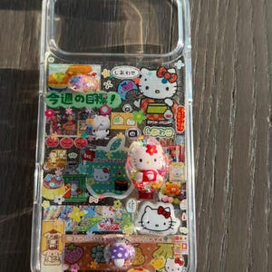 Op de afbeelding: Een doorzichtig telefoonhoesje versierd met een collage van kleurrijke stickers en driedimensionale bedels. Het ontwerp bevat meerdere Hello Kitty-illustraties, Japanse tekst waaronder "今週の目標!", diverse voedselpictogrammen en kleine glinsterende accenten die over het hele oppervlak zijn verwerkt.