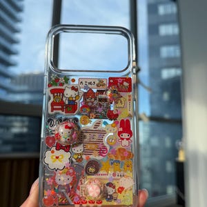 Op de afbeelding: Een doorzichtig telefoonhoesje versierd met een collage van kleurrijke stickers en kleine driedimensionale bedels met Hello Kitty, My Melody en diverse etenswaren. Het ontwerp bevat Japanse tekst, glitteraccenten en kleine figuurtjes.
