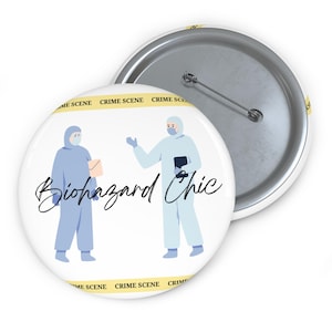 Puede incluir: Un botón blanco con un alfiler plateado. Presenta la inscripción "Biohazard Chic" y una ilustración de dos figuras con trajes de protección azul claro. Las palabras "CRIME SCENE" están impresas alrededor del borde del botón.