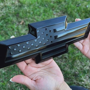 May include: A glossy black car emblem featuring a stylized American flag design with stars and stripes. The emblem is held in front of a green grassy background.