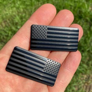 May include: Two black rectangular emblems with a raised American flag design. The flag features a field of stars and stripes. The emblems are held in a hand against a background of green grass.