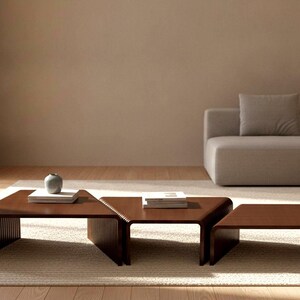 May include: A set of three dark brown wooden coffee tables with curved, ribbed sides. The tables are arranged in a stepped formation, with a small gray vase and a stack of books placed on the left table and a stack of books on the center table.