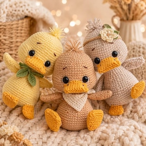 May include: Three handmade crochet ducklings in shades of yellow, tan, and beige. Each duckling has a yellow beak and feet, with black eyes. One duckling has a green leaf accent, another a headband with a flower, and the third a white bandana. They are sitting on a textured, cream-colored surface.