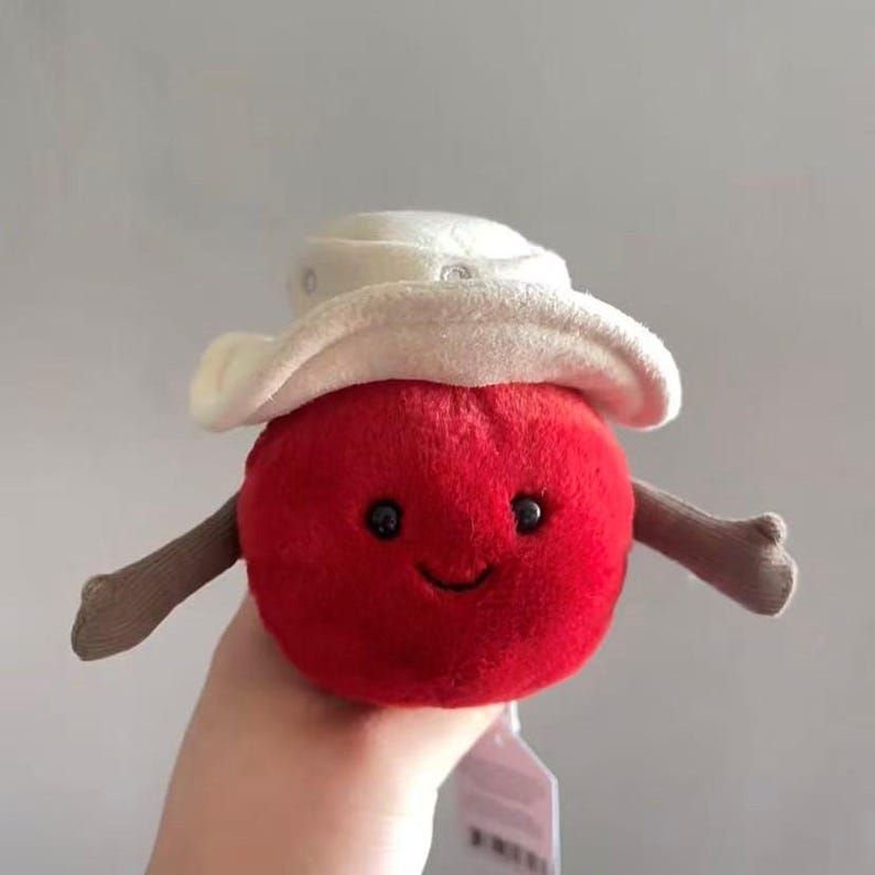 Puede incluir: Un juguete de peluche rojo con forma de manzana con un sombrero de vaquero blanco, brazos peque&ntilde;os y una cara sonriente. El juguete blando est&aacute; dise&ntilde;ado con una expresi&oacute;n amigable y es adecuado para ni&ntilde;os.