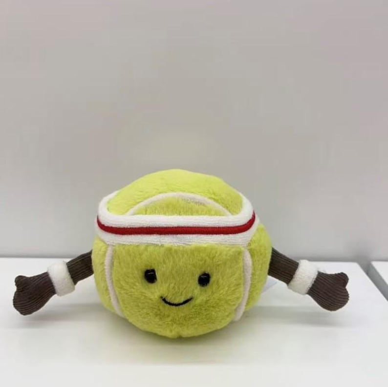 Puede incluir: Un juguete de peluche con forma de pelota de tenis con una cara sonriente, que lleva una diadema blanca y roja y brazos marrones con mu&ntilde;equeras blancas. La pelota de tenis es de color amarillo-verdoso brillante.