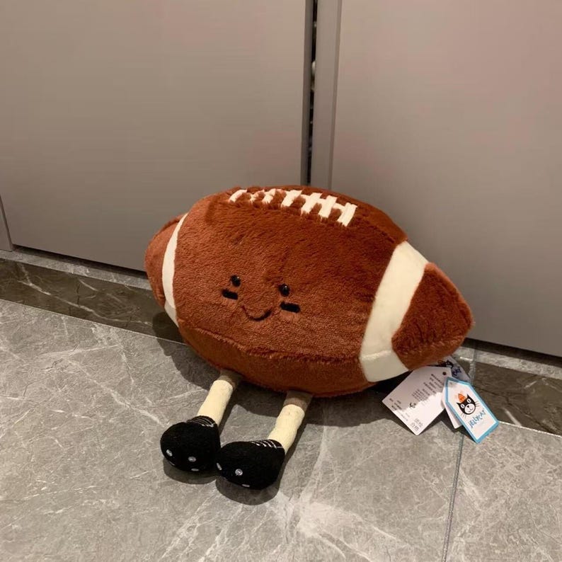 Puede incluir: Un bal&oacute;n de f&uacute;tbol americano de peluche marr&oacute;n con rayas y costuras blancas, con una cara sonriente. Tiene patas beige con zapatos de lunares blancos y negros. Una etiqueta est&aacute; adjunta.