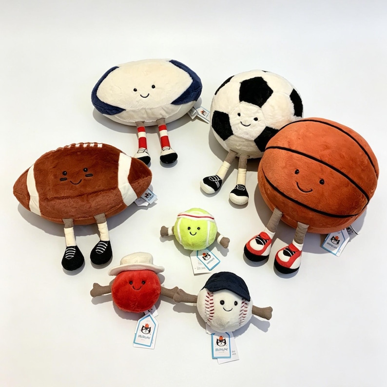 Puede incluir: Una colecci&oacute;n de peluches de balones deportivos, incluyendo un bal&oacute;n de f&uacute;tbol americano, un bal&oacute;n de f&uacute;tbol, un bal&oacute;n de baloncesto, un bal&oacute;n de b&eacute;isbol, una pelota de tenis y un bal&oacute;n de rugby. Cada juguete tiene una cara sonriente, piernas y zapatos.
