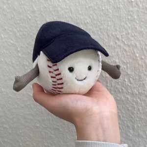 Puede incluir: Un juguete de b&eacute;isbol de peluche con una cara sonriente, costuras rojas y una gorra de b&eacute;isbol azul marino. El juguete tiene brazos grises y est&aacute; dise&ntilde;ado para parecer un personaje amigable. El b&eacute;isbol se sostiene en una mano.
