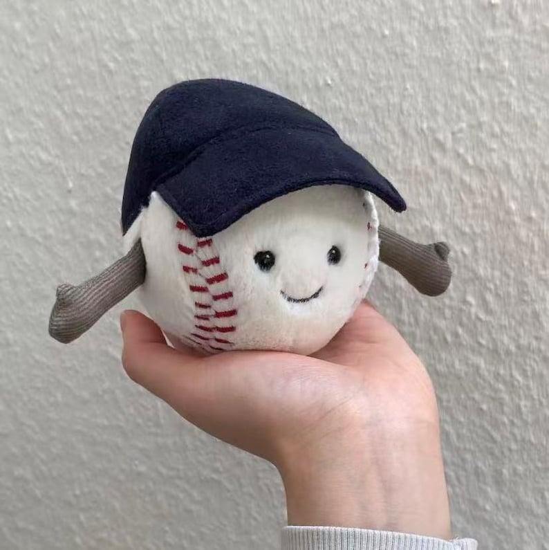 Puede incluir: Un juguete de b&eacute;isbol de peluche con una cara sonriente, costuras rojas y una gorra de b&eacute;isbol azul marino. El juguete tiene brazos grises y est&aacute; dise&ntilde;ado para parecer un personaje amigable. El b&eacute;isbol se sostiene en una mano.