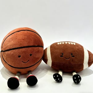 Puede incluir: Dos pelotas deportivas de peluche: una de baloncesto y otra de f&uacute;tbol americano. La de baloncesto es naranja con l&iacute;neas negras, y la de f&uacute;tbol americano es marr&oacute;n con rayas blancas. Ambas tienen caras sonrientes, pies negros y detalles rojos y blancos.