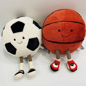 Puede incluir: Dos balones deportivos de peluche: un bal&oacute;n de f&uacute;tbol y un bal&oacute;n de baloncesto. El bal&oacute;n de f&uacute;tbol es blanco con pent&aacute;gonos negros, y el bal&oacute;n de baloncesto es naranja con l&iacute;neas negras. Ambos tienen caras sonrientes, piernas marrones y llevan zapatos.