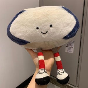 Puede incluir: Un peluche con forma de nube con una cara sonriente. La nube es de color crema con un borde azul marino. Tiene dos piernas con calcetines a rayas rojas y blancas y zapatos blancos y negros.