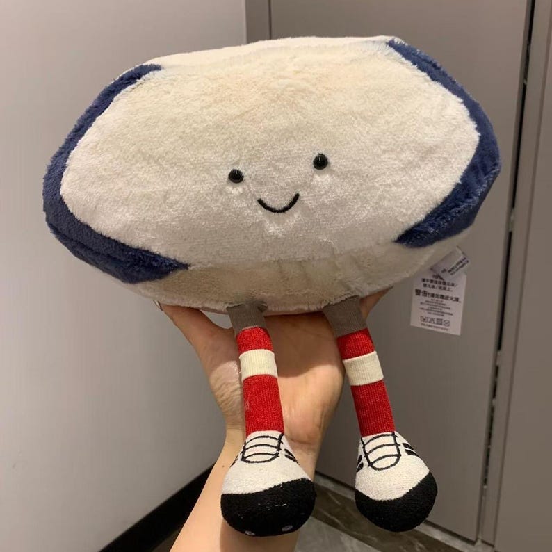 Puede incluir: Un peluche con forma de nube con una cara sonriente. La nube es de color crema con un borde azul marino. Tiene dos piernas con calcetines a rayas rojas y blancas y zapatos blancos y negros.