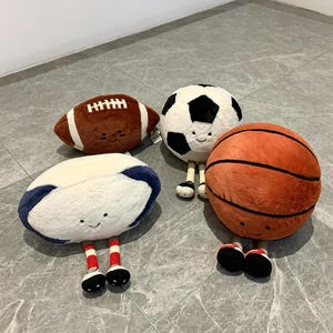 Puede incluir: Cuatro pelotas deportivas de peluche: un bal&oacute;n de f&uacute;tbol americano marr&oacute;n, un bal&oacute;n de f&uacute;tbol blanco y negro, un bal&oacute;n de baloncesto naranja y un bal&oacute;n de rugby blanco y azul marino. Cada uno tiene una cara sonriente y piernas con zapatos.