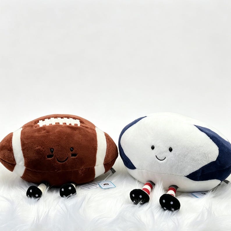 Puede incluir: Dos juguetes de peluche con forma de bal&oacute;n deportivo. Uno es un bal&oacute;n de f&uacute;tbol americano marr&oacute;n con rayas blancas y el otro es un bal&oacute;n de rugby blanco y azul marino. Ambos tienen caras sonrientes y pies blancos y negros.