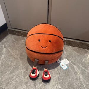 Puede incluir: Un peluche de baloncesto naranja con rayas negras y una cara sonriente. Tiene piernas grises y lleva zapatillas rojas y blancas. Una etiqueta est&aacute; adjunta al juguete.